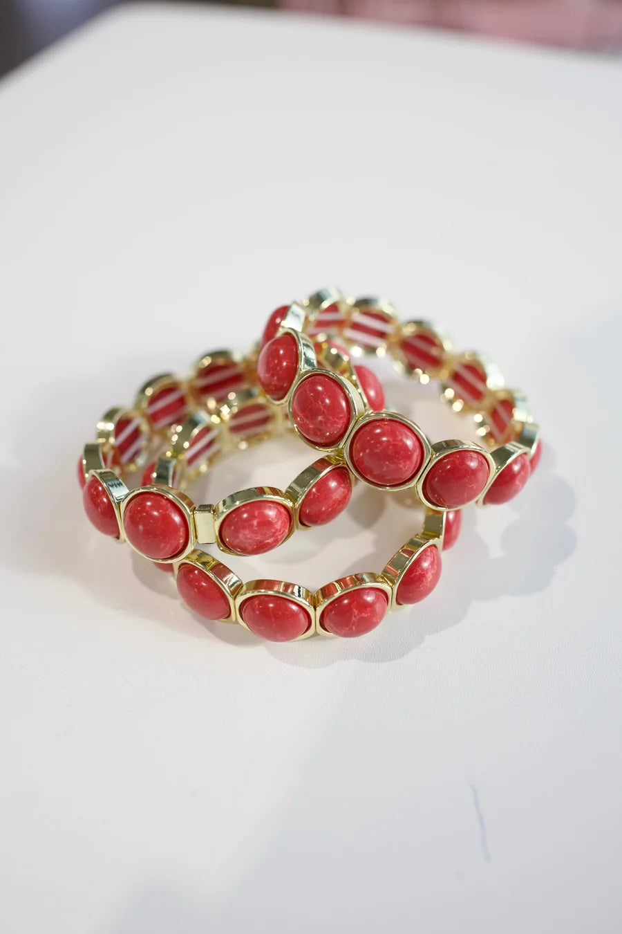 A stack of three gold-plated stretch bracelets, each featuring a row of oval red cabochon stones, displayed on a white surface.