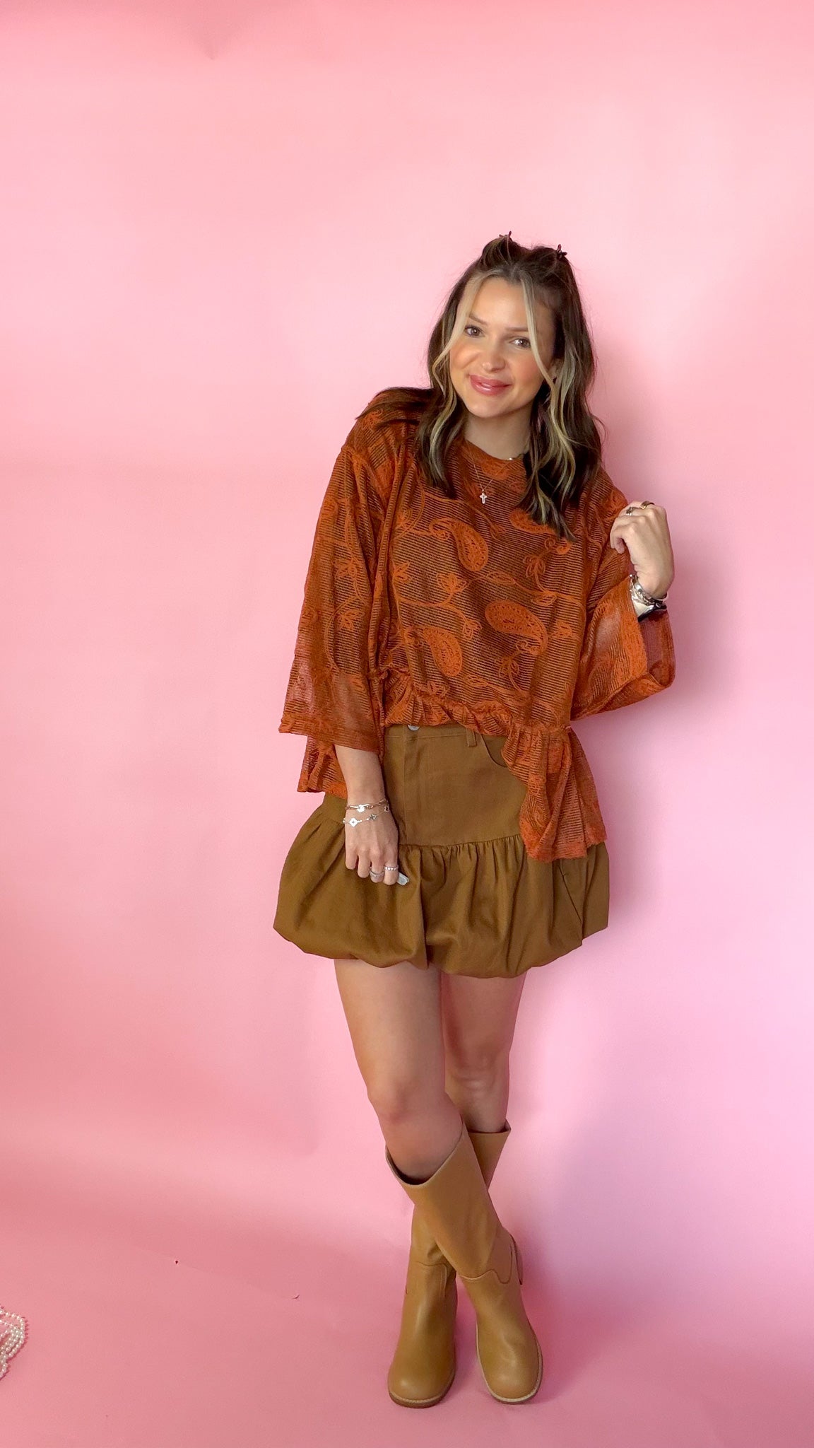 A woman modeling a rust-orange patterned long-sleeve top and brown ruffled mini skirt with tan knee-high boots, standing against a solid pink backdrop.