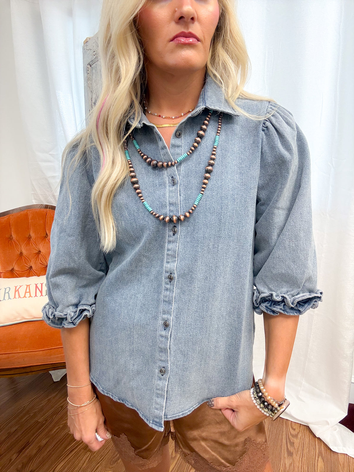 A woman wearing a light-wash blue denim button-up shirt with ruffled sleeves, layered with multiple beaded necklaces, standing in front of a white curtain backdrop.