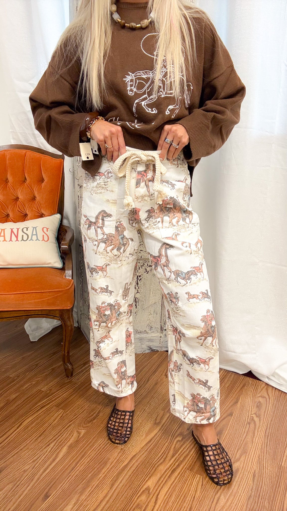 A person wearing loose-fit cream pants with a brown horse-and-rider print, styled with a brown sweatshirt and brown woven shoes, standing in a room with a wooden floor, white curtain backdrop, and an orange chair.