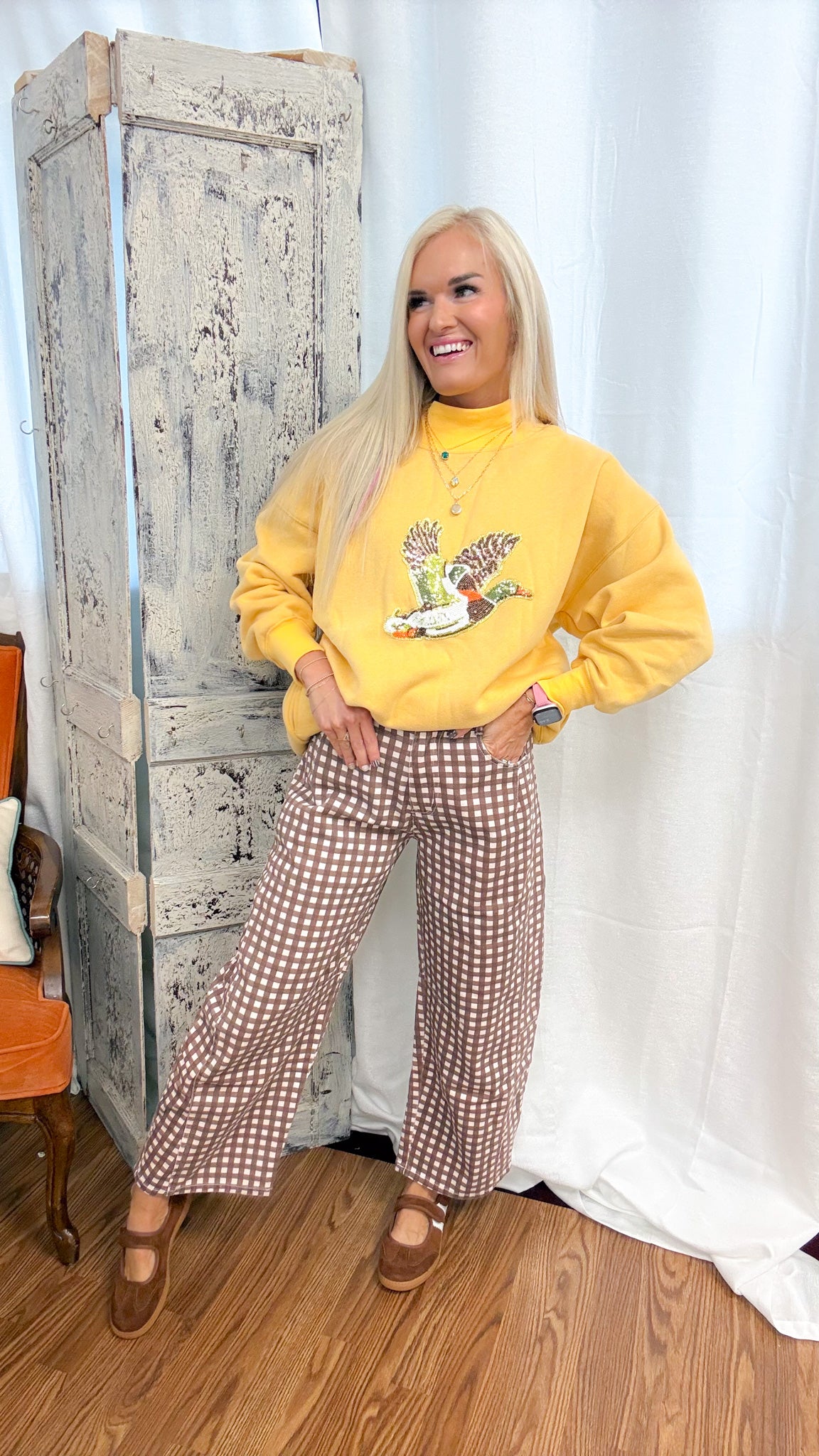 A woman posing in a yellow mock-neck sweatshirt with a duck graphic and brown-and-white gingham wide-leg pants, standing in front of a rustic white curtain and a distressed wooden cabinet.