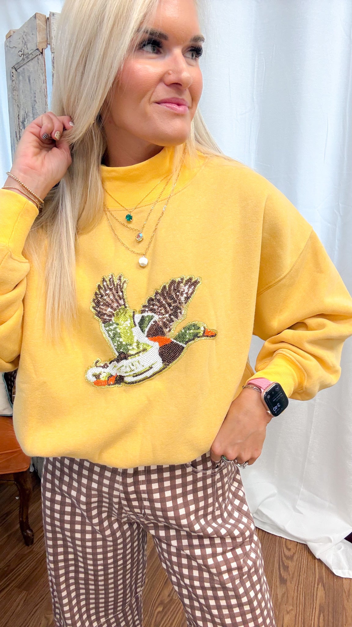 A woman wearing a yellow sweatshirt with a large embroidered duck graphic on the front, paired with brown and white gingham pants.