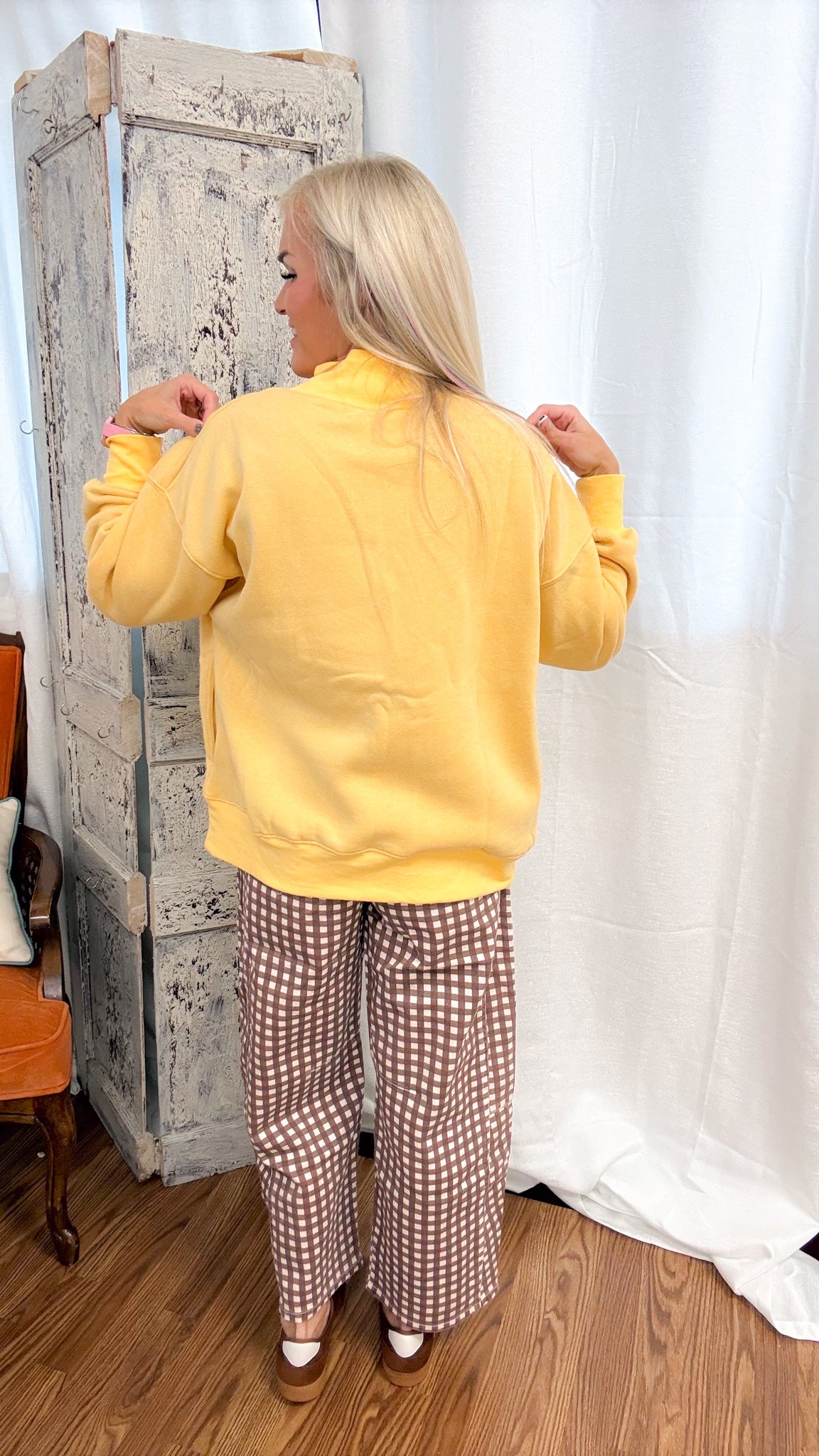 A woman with blonde hair is shown from behind wearing a yellow sweatshirt and brown-and-white checkered pants, standing in a room with a rustic wooden cabinet and a white curtain backdrop.