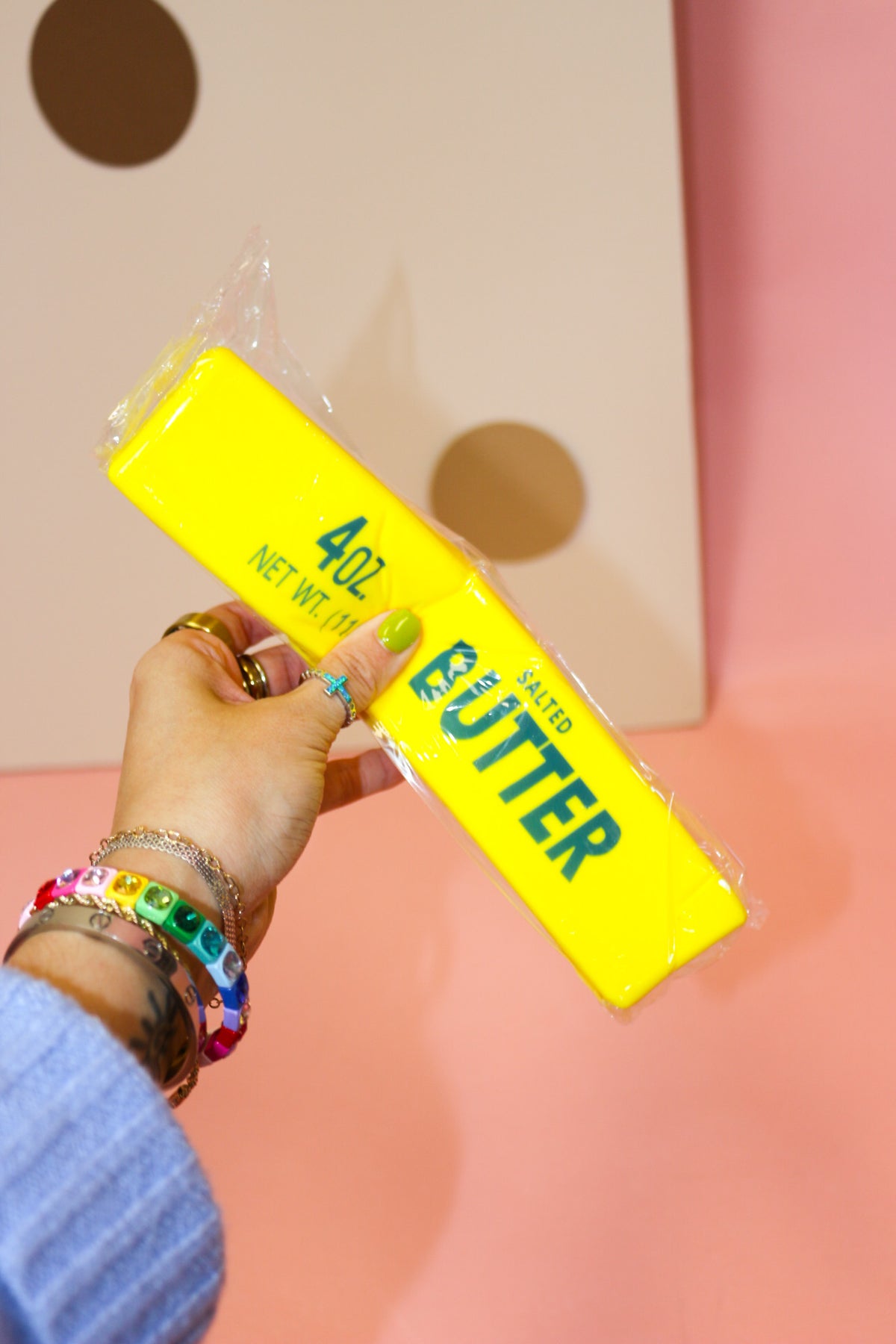Hand holding a rectangular yellow butter squishy with green butter-label graphics against a pink and dotted backdrop.