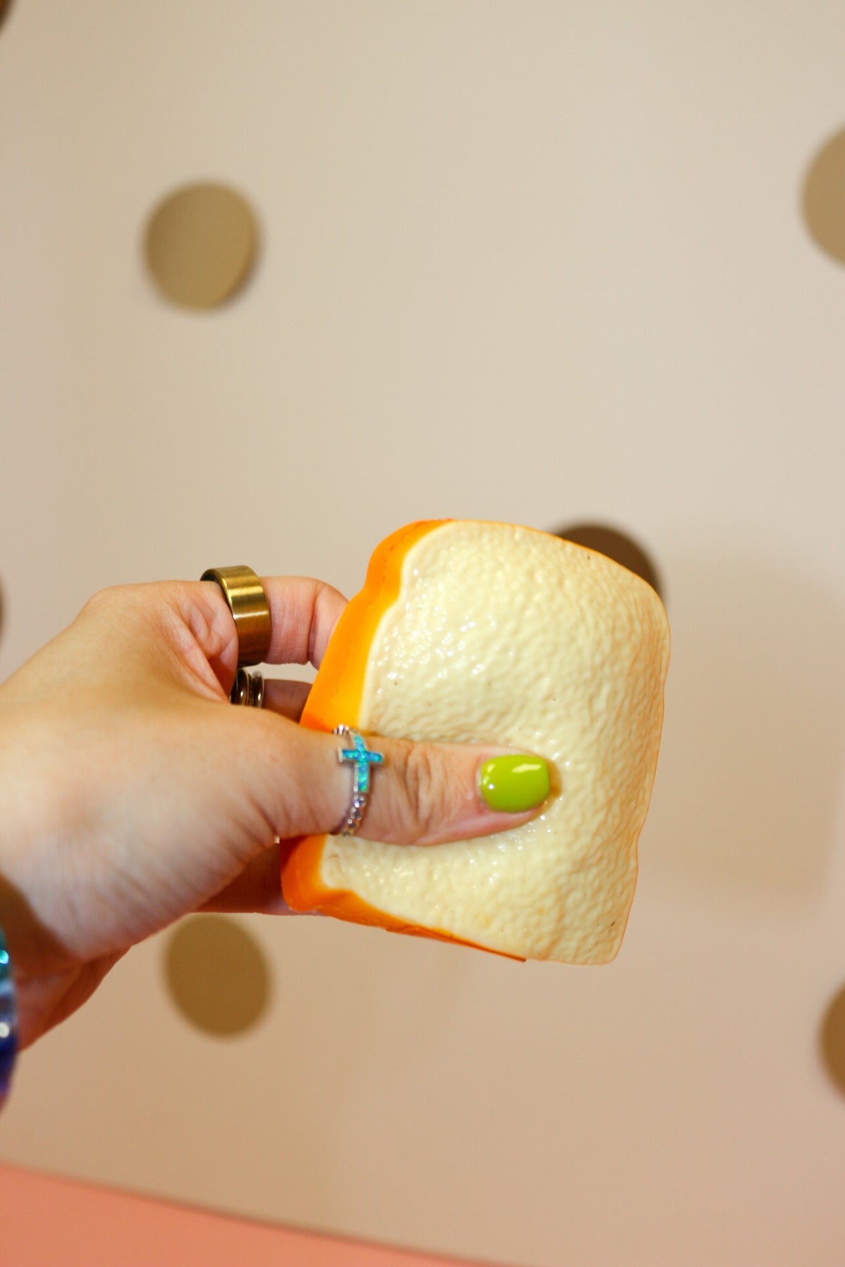 Hand squeezing a cream-colored toast squishy with golden-orange edging against a beige dotted backdrop.