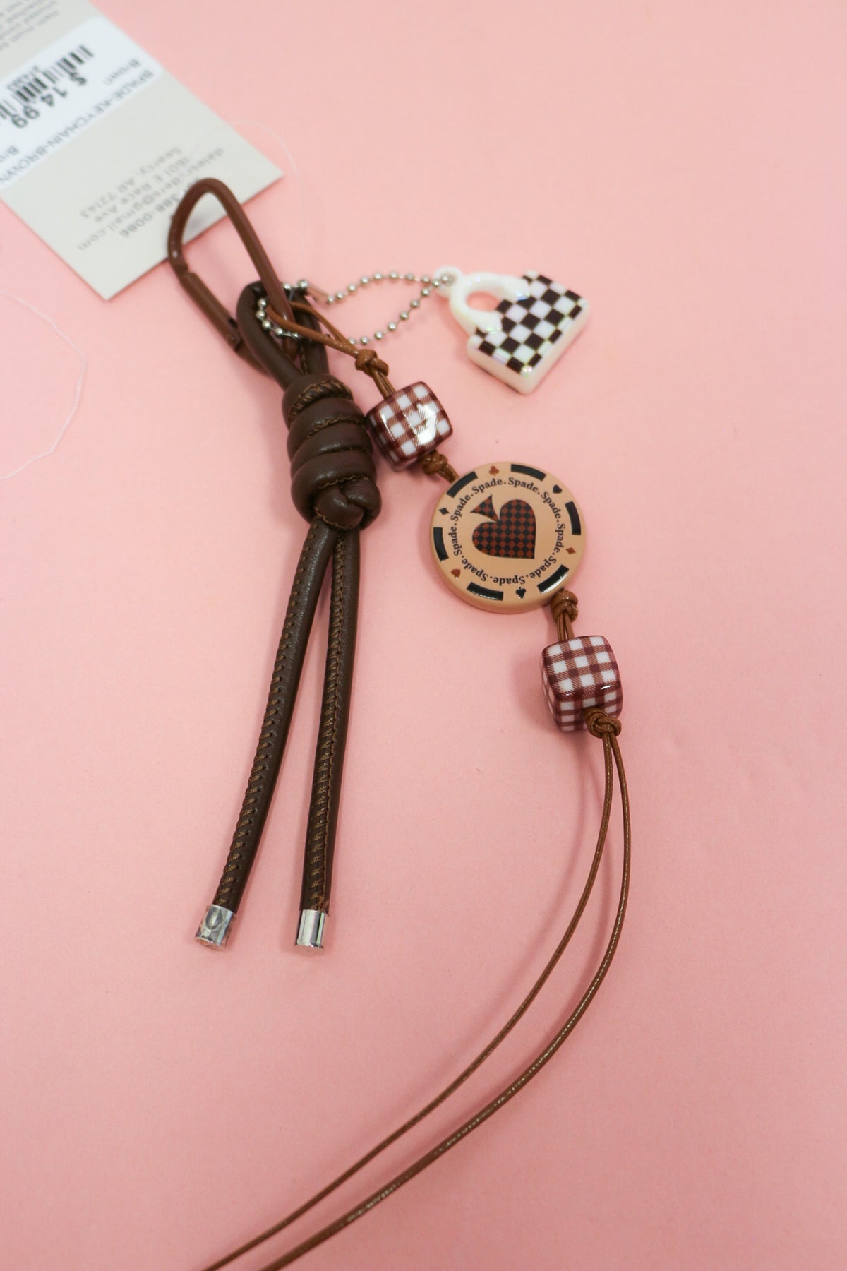 Brown Suit Of Spade keychain with knotted cords, gingham beads, spade disc and checkerboard handbag charm on pink.