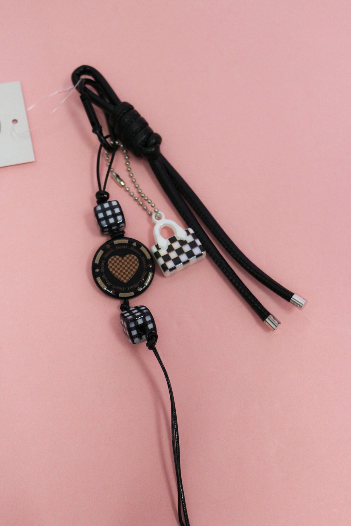 Black heart keychain charm with checkered beads, mini handbag charm and knotted cords on a pink background.