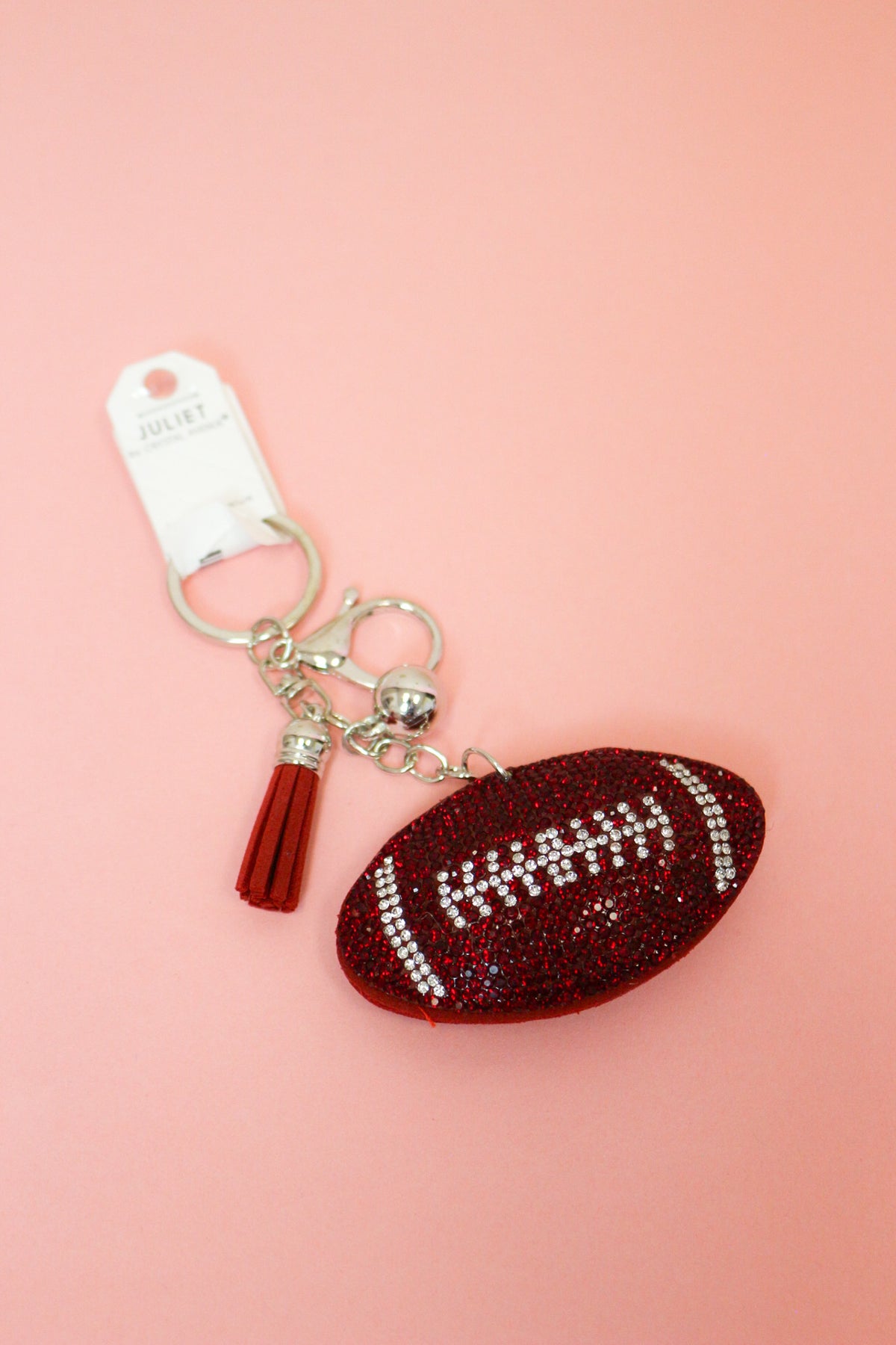 Red rhinestone football keychain charm with matching tassel, silver-tone key ring and clasp shown on a pink background.