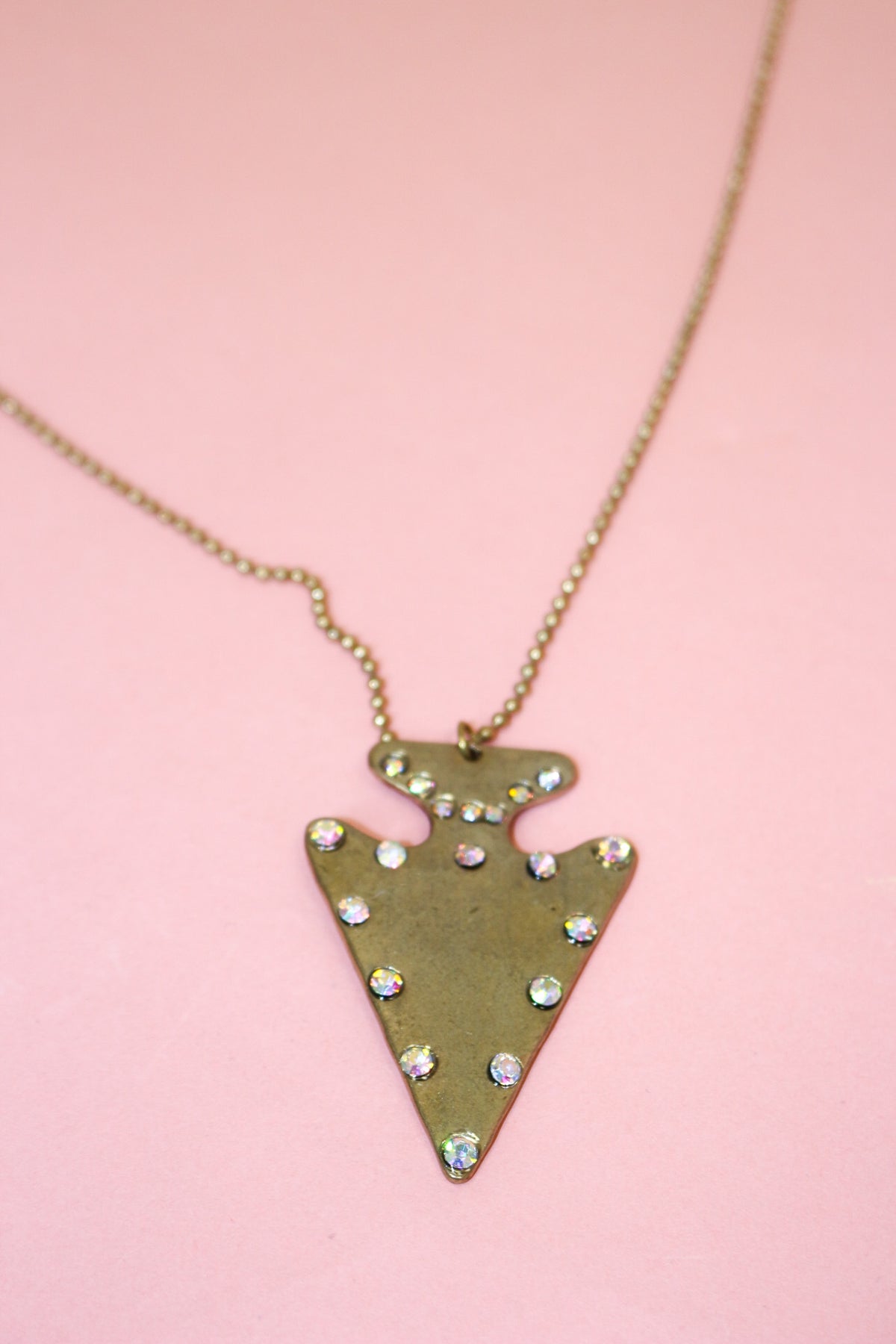 Close-up of antiqued gold-tone arrowhead pendant with scattered round iridescent jewel accents on a fine-link chain.