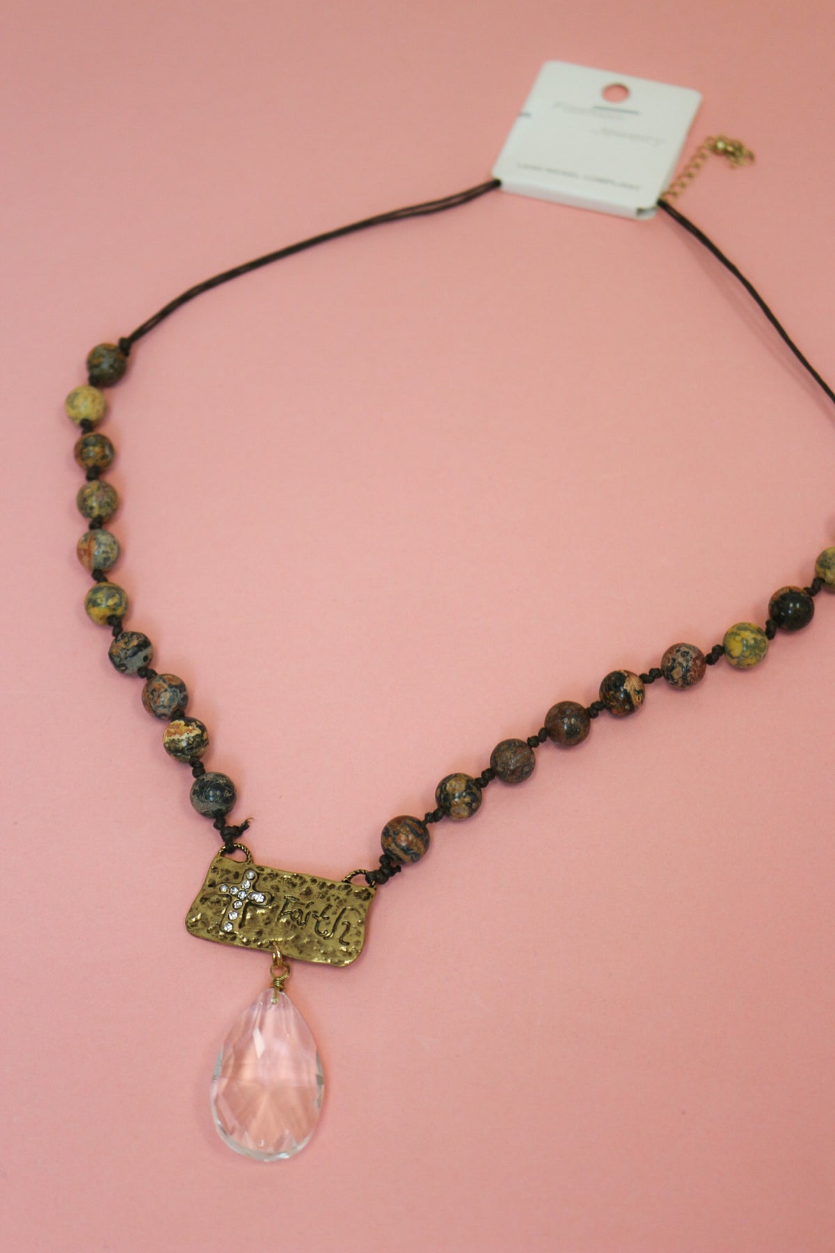 Brown and earthy mixed-tone beaded necklace on dark cord with faith plaque and clear teardrop pendant on pink background.