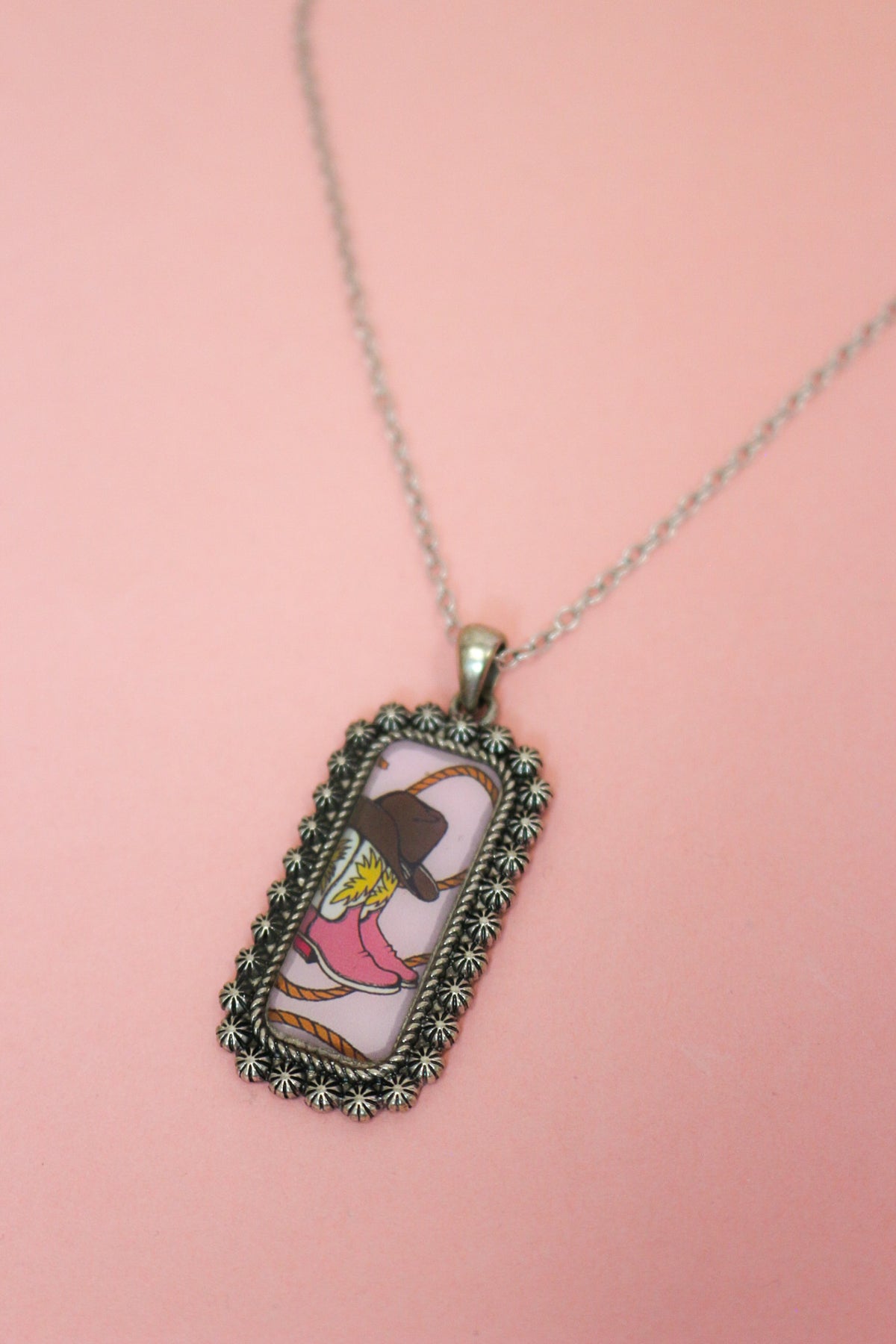 Close-up of Western tag pendant with pink cowboy boot, brown hat, rope illustration, and ornate silver-tone border.