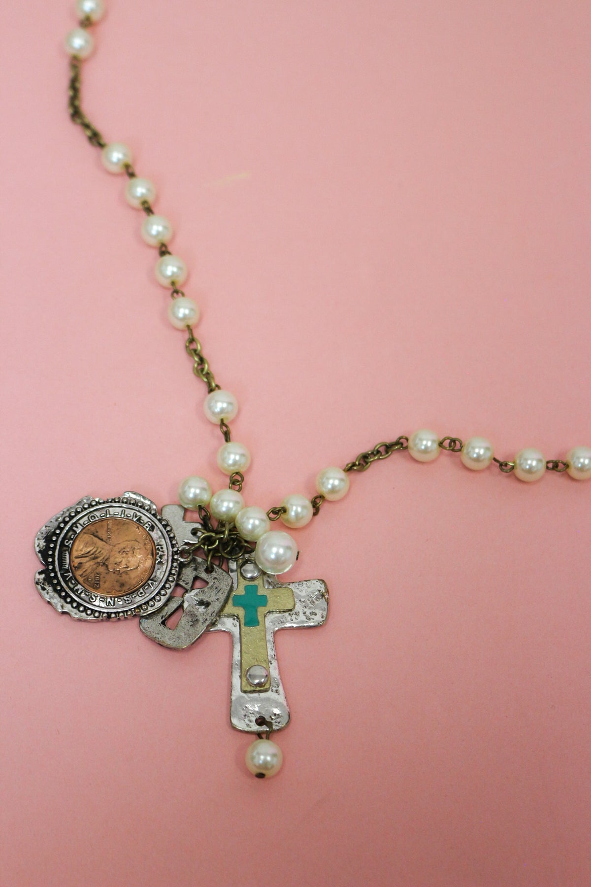 Close-up of pearl-look Western necklace with penny charm, textured cross pendant and turquoise-colored accent on pink.