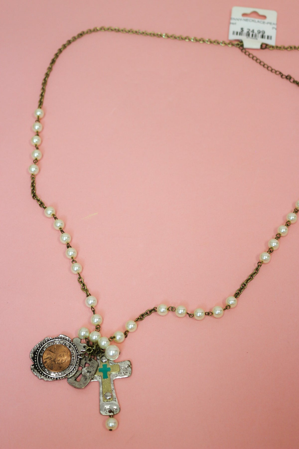 Pearl-look beaded chain Western necklace with penny charm and layered cross pendant laid out on a pink background.