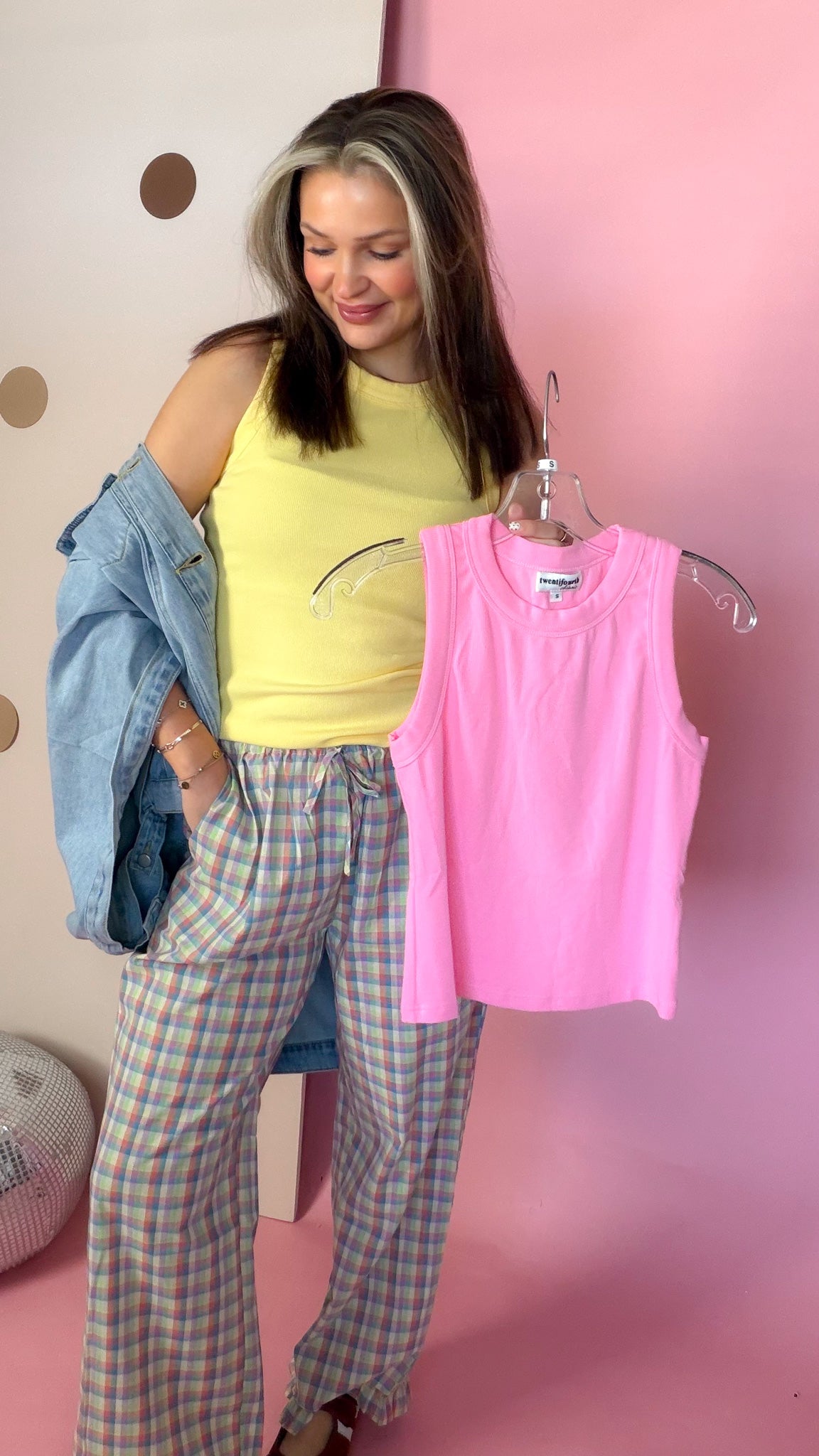 Model wearing Daisy yellow ribbed tank top and holding a pink sleeveless top on a hanger.