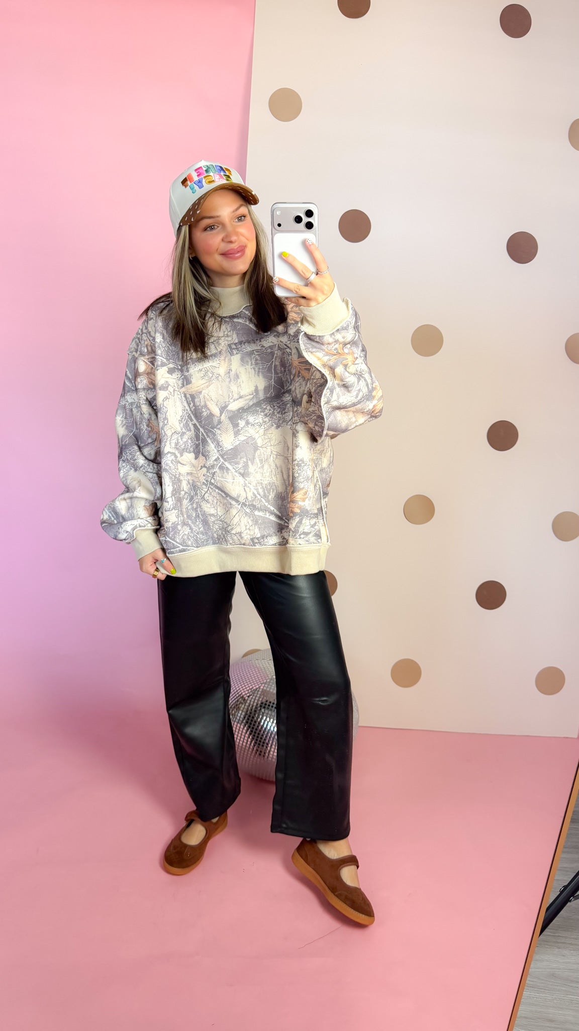 Full-length mirror view of women's taupe camo sweatshirt with black pants, brown flats, and a cap.