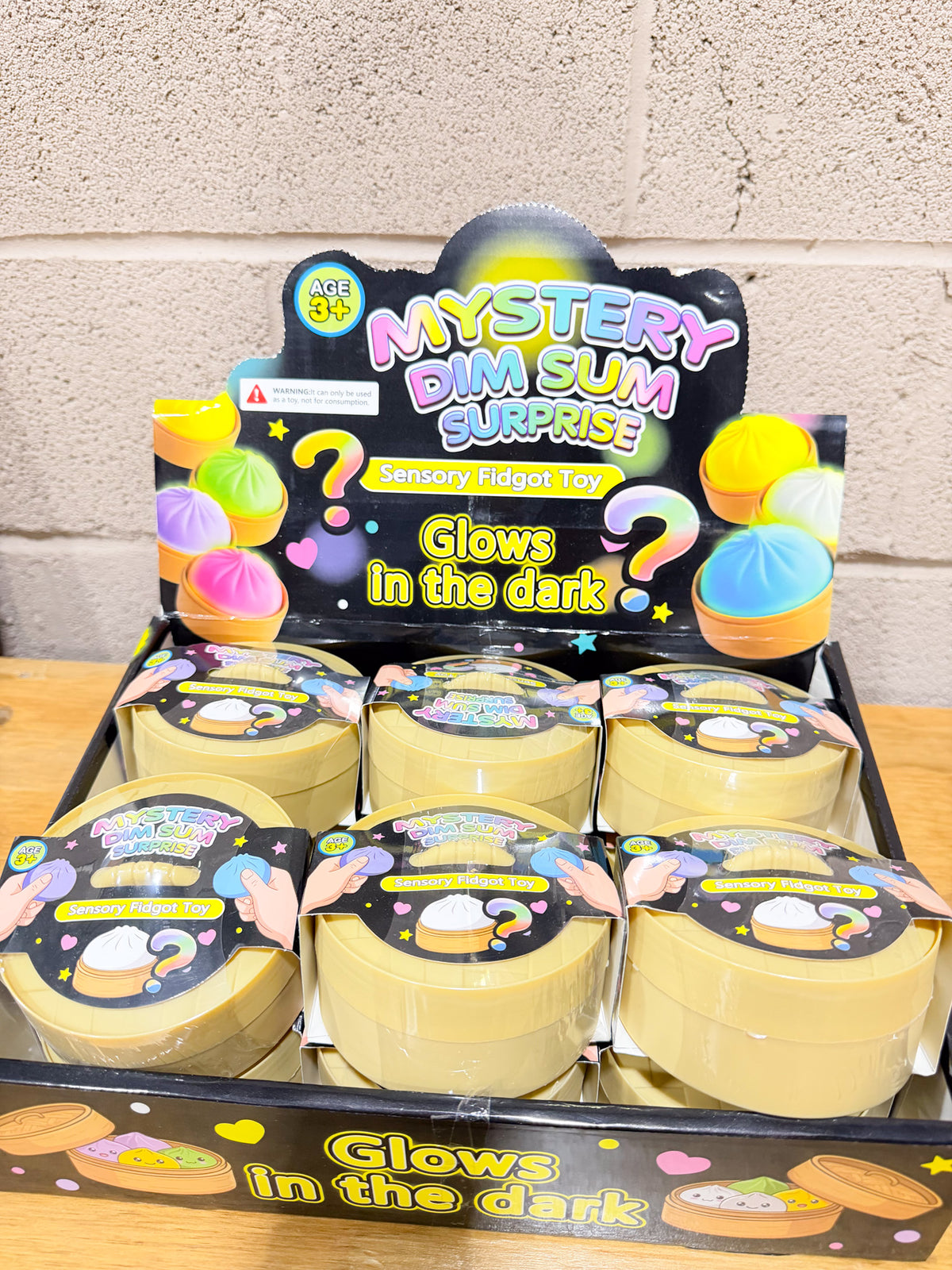 Full display of glow-in-the-dark dumpling squishy packages in yellow steamer-style containers on a wooden tabletop.