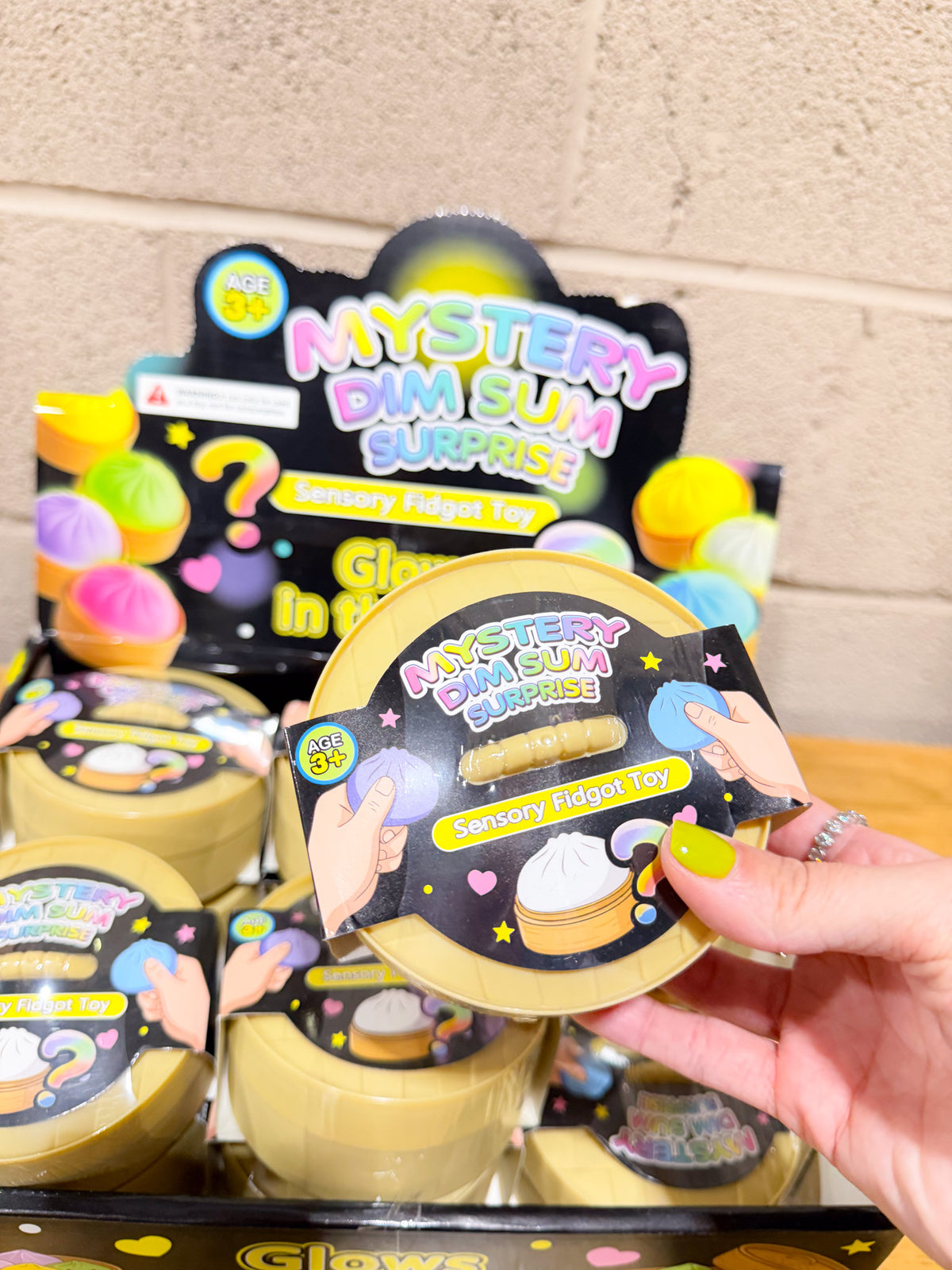 Hand holding a round yellow Mystery Dim Sum Surprise package labeled Sensory Fidget Toy and ages 3+.