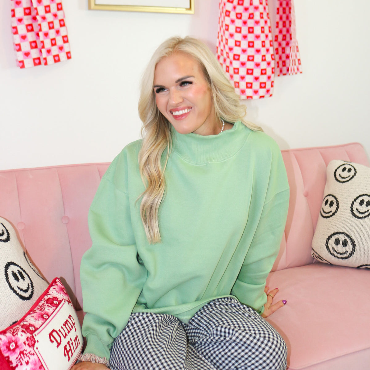Woman in a green sweater and checkered pants standing in a room with pink couch, decorative pillows, and wall art.