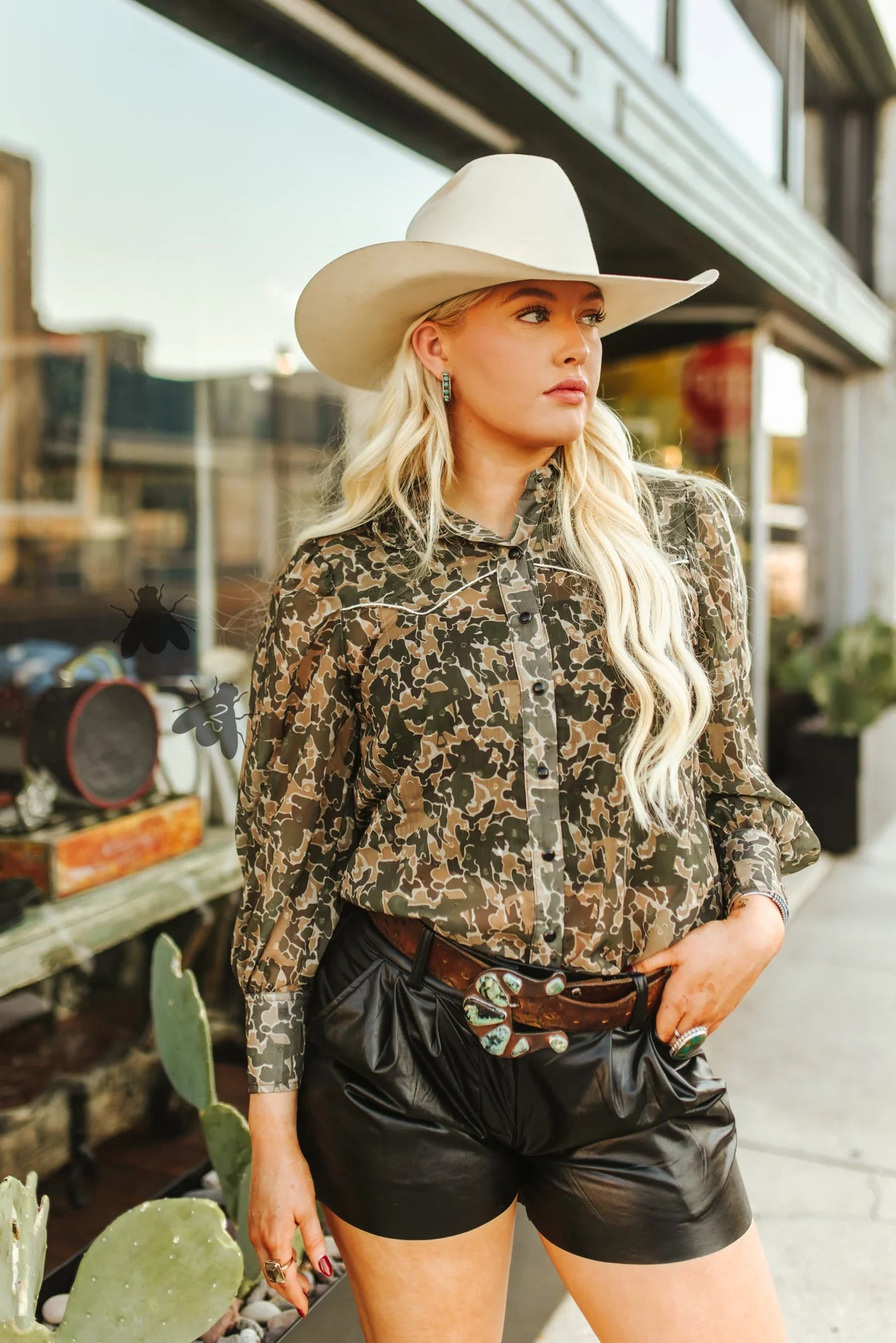 Woman wearing a camouflage shirt, black leather shorts, and a cowboy hat outdoors.