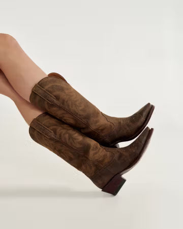 Brown suede boots worn by a person on a light gray background. Tecovas women’s cowboy boots