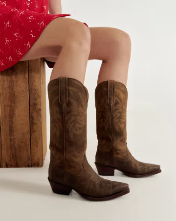Brown cowboy boots worn by a person sitting on a wooden stool with a white background. Tecovas women’s cowboy boots