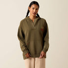 A woman with dark braided hair wears an olive green pullover hoodie with a unique split neckline and a small embroidered graphic on the left chest, paired with beige pants, against a solid light beige background.