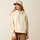 A woman wearing a cream-colored cropped hoodie with a drawstring hood, paired with brown pants and a matching cream baseball cap, photographed against a solid light beige background.
