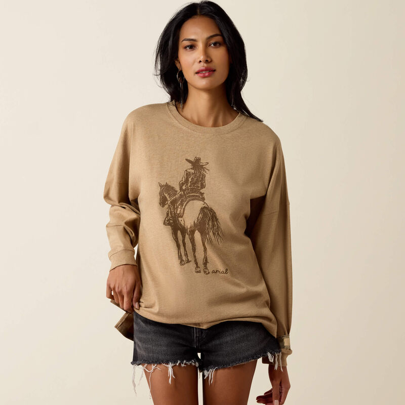 A woman modeling a beige crewneck sweatshirt with a brown graphic of a cowboy on a horse, paired with denim shorts, against a solid light beige background.