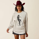 A woman modeling a cream-colored long-sleeve crewneck sweatshirt with a black cowboy graphic print, styled with a dark red cowboy hat and denim shorts against a solid light beige background.