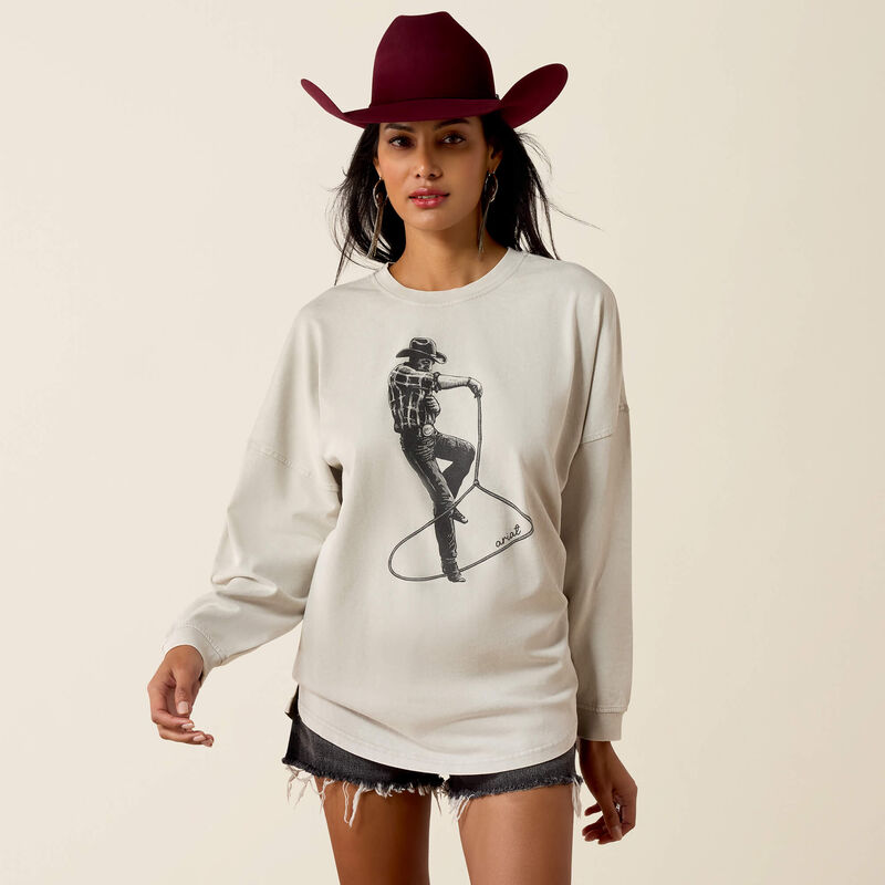 A woman modeling a cream-colored long-sleeve crewneck sweatshirt with a black cowboy graphic print, styled with a dark red cowboy hat and denim shorts against a solid light beige background.