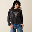 A woman modeling a dark charcoal gray long-sleeve t-shirt with a white line art graphic of a longhorn steer skull on the front, paired with blue jeans against a solid light beige background.
