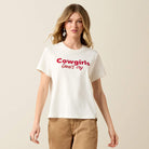 A woman modeling a white short-sleeve t-shirt with 'Cowgirls Don't cry' printed in red across the chest, paired with tan pants, against a solid light beige background.