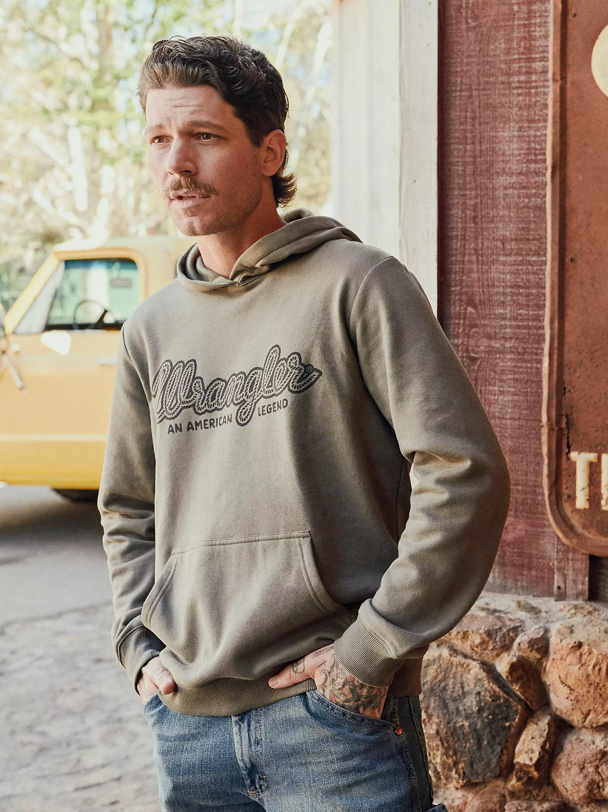 Wrangler Legacy script graphic hoodie in olive, worn by man outdoors by a truck