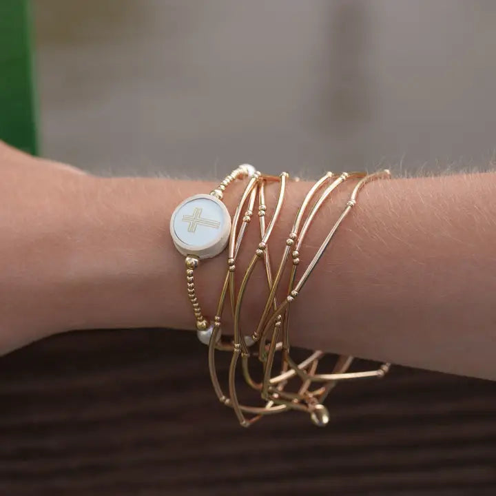 Beloved Tap To Pray gold-tone beaded cross bracelet worn on a wrist with layered gold-tone bracelets.