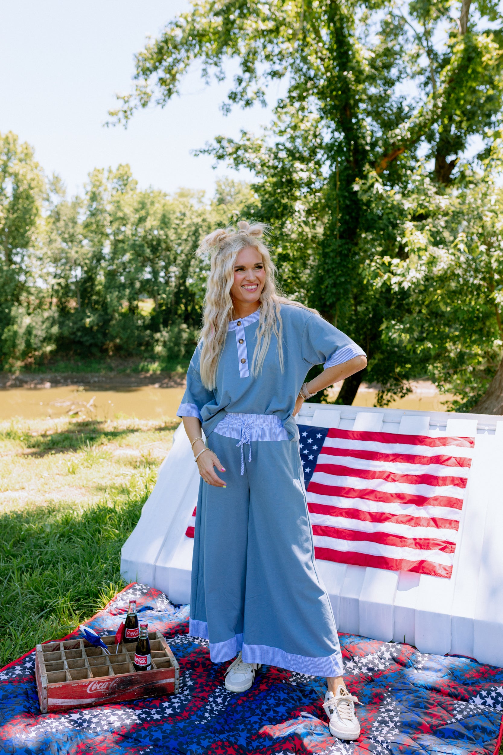 Woman in a blue outfit standing outdoors with an American flag and picnic blanket.