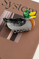 A duck-shaped hair claw clip with a glossy green head, yellow beak, orange body, and black-and-white patterned wing, attached to a gray clip base, displayed on a brown textured surface.