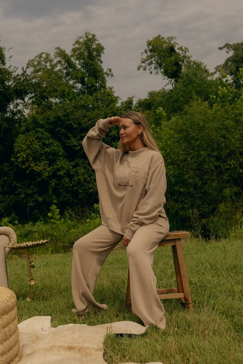 A woman wearing a beige lounge set sits on a wooden stool outdoors on a grassy lawn with trees in the background. The set includes a sweatshirt and matching wide-leg pants in a light taupe-beige color.