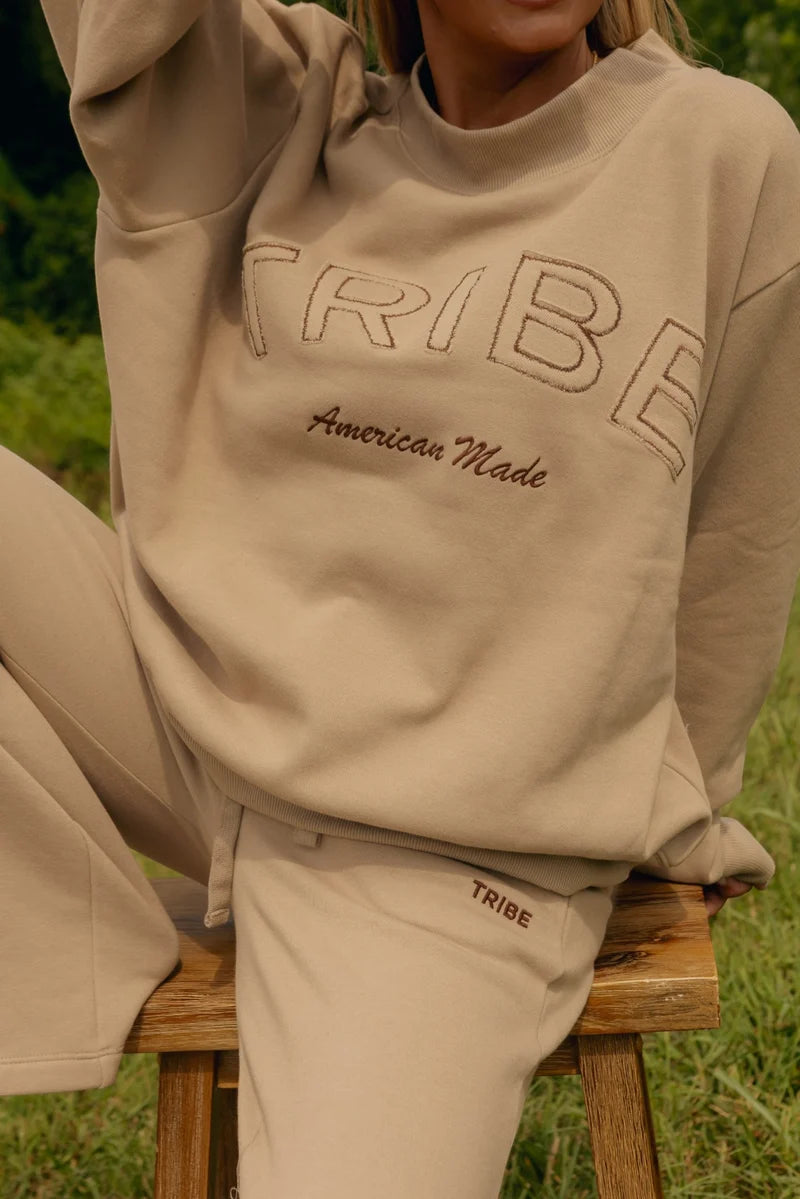 A person wearing a beige mockneck sweatshirt and matching sweatpants, sitting on a wooden stool outdoors. The sweatshirt features 'TRIBE' embroidered across the chest in a large outlined font with 'American Made' embroidered underneath.