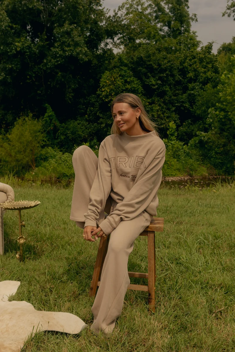 A woman wearing a beige lounge set consisting of a sweatshirt and matching sweatpants sits on a wooden stool in a grassy outdoor setting with trees in the background.