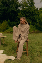 A woman wearing a beige lounge set consisting of a sweatshirt and matching sweatpants sits on a wooden stool in a grassy outdoor setting with trees in the background.