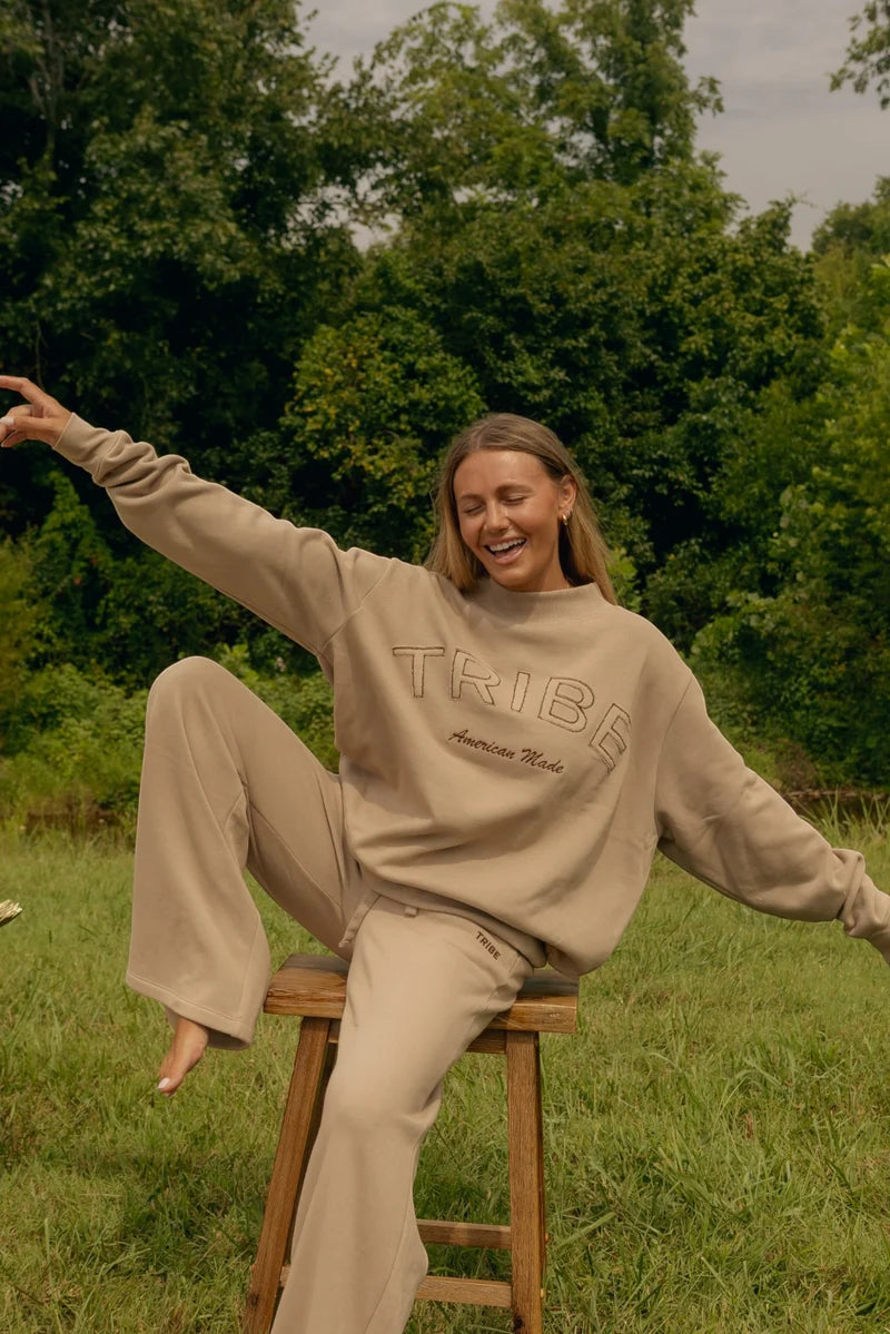 A woman wearing a beige matching sweatshirt and sweatpants lounge set sits on a wooden stool in a grassy outdoor setting with trees in the background. The sweatshirt features the word 'TRIBE' and 'American Tribe' printed on the front.