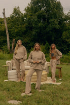 Three women modeling matching beige lounge sets in an outdoor grassy setting with a sofa and trees in the background.