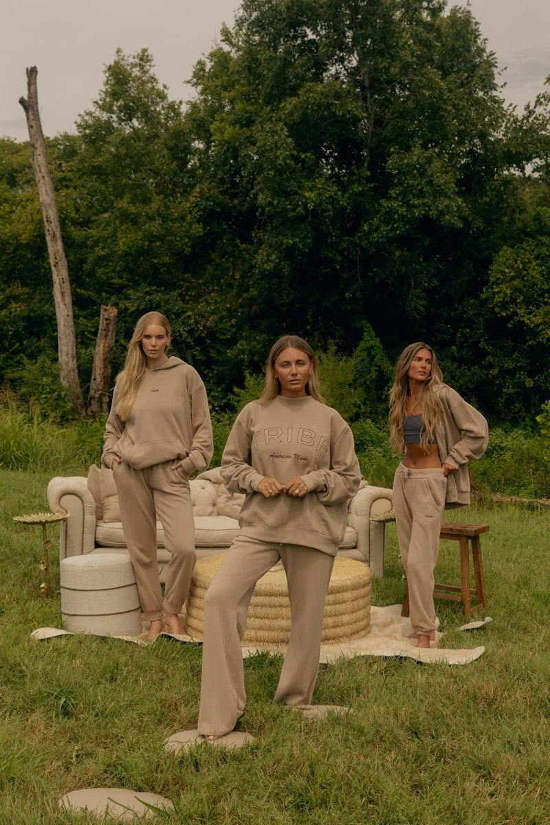 Three women modeling matching beige lounge sets in an outdoor grassy setting with a sofa and trees in the background.