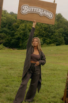 A woman stands in a grassy outdoor field holding up a wooden sign that reads 'BUTTERLAND' in raised letters. She is wearing a dark brown cropped tank top, matching dark brown sweatpants, and a long dark open-front coat.
