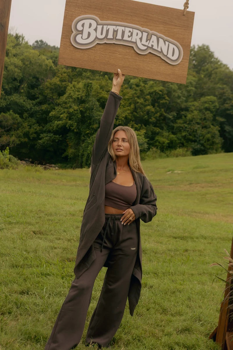 A woman stands in a grassy outdoor field holding up a wooden sign that reads 'BUTTERLAND' in raised letters. She is wearing a dark brown cropped tank top, matching dark brown sweatpants, and a long dark open-front coat.