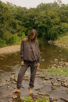 A woman stands barefoot in a shallow rocky stream surrounded by dense green trees, wearing a dark brown sweatshirt and matching sweatpants with a tonal graphic on the chest.