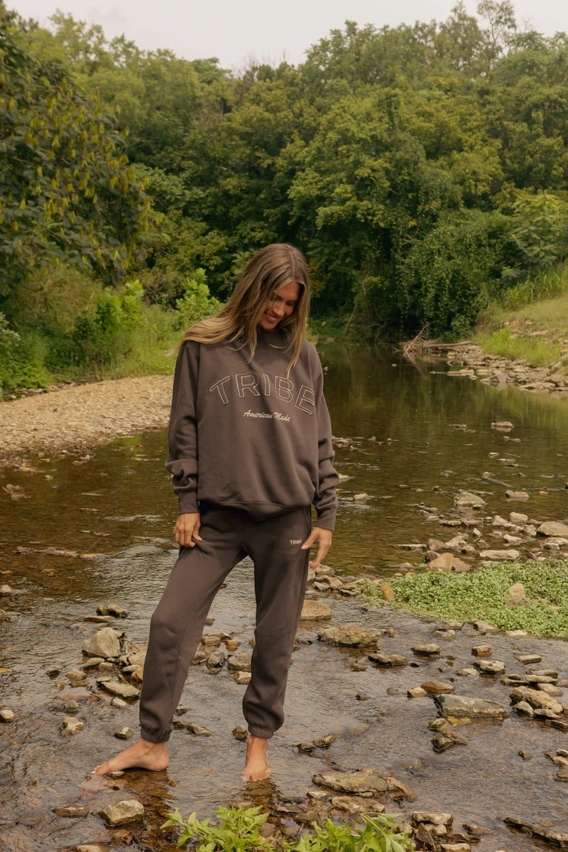 A woman stands barefoot in a shallow rocky stream surrounded by dense green trees, wearing a dark brown sweatshirt and matching sweatpants with a tonal graphic on the chest.