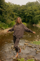 A woman stands barefoot in a shallow rocky river, wearing a dark brown ribe sweatshirt and matching sweatpants with white text. The background is a lush green forest.