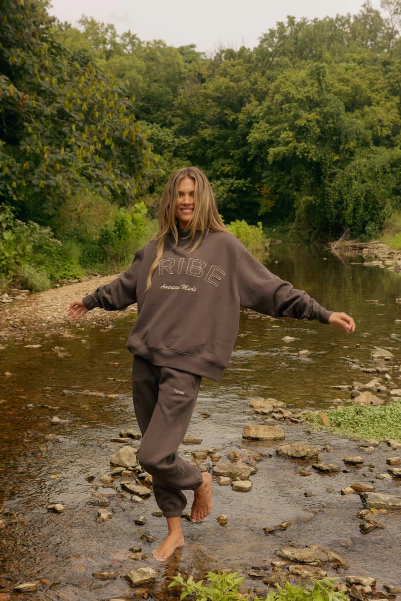 A woman stands barefoot in a shallow rocky river, wearing a dark brown ribe sweatshirt and matching sweatpants with white text. The background is a lush green forest.