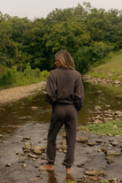 A woman stands barefoot in a shallow rocky stream, viewed from behind, wearing a dark brown matching lounge set consisting of a long-sleeve top and sweatpants, with a lush green forest in the background.